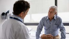 A patient sits on an exam table and listens to the doctor in the foreground talking